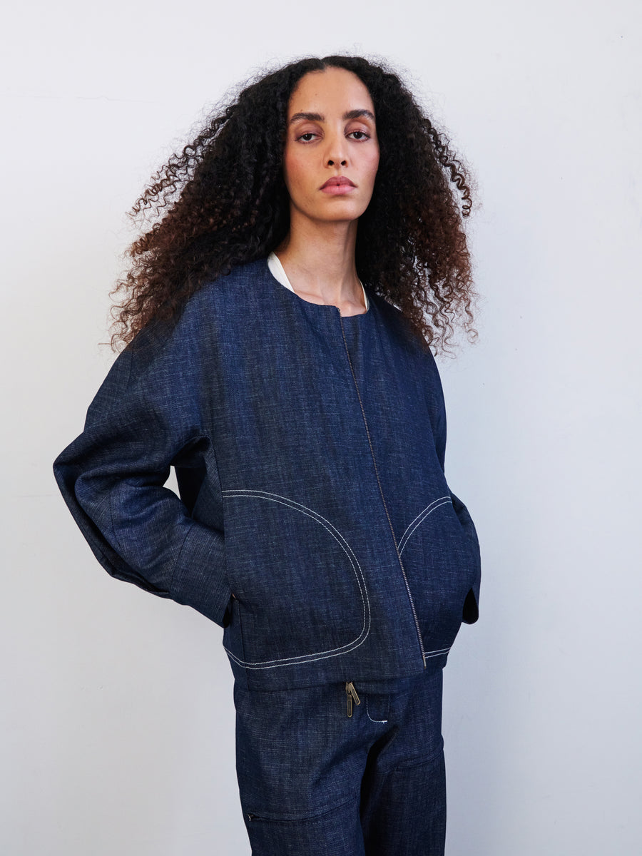 A person with long, curly hair wears a blue Akeo Bomber Jacket by Zero + Maria Cornejo, standing against a plain white background.