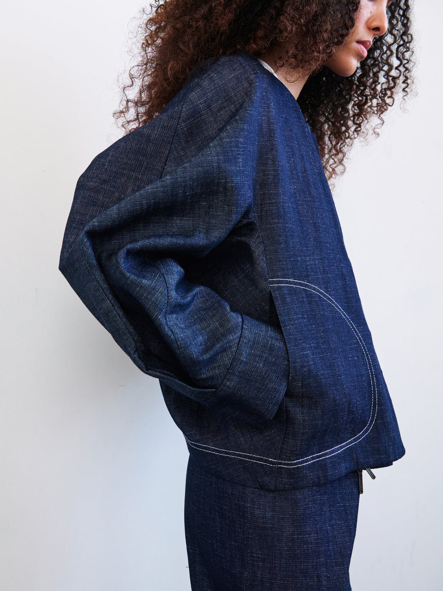 A person with curly hair in a dark blue Akeo Bomber Jacket by Zero + Maria Cornejo, made from GOTS-certified cotton, is shown from the side against a plain white background. The New York-made jacket features visible white stitching on the pocket.