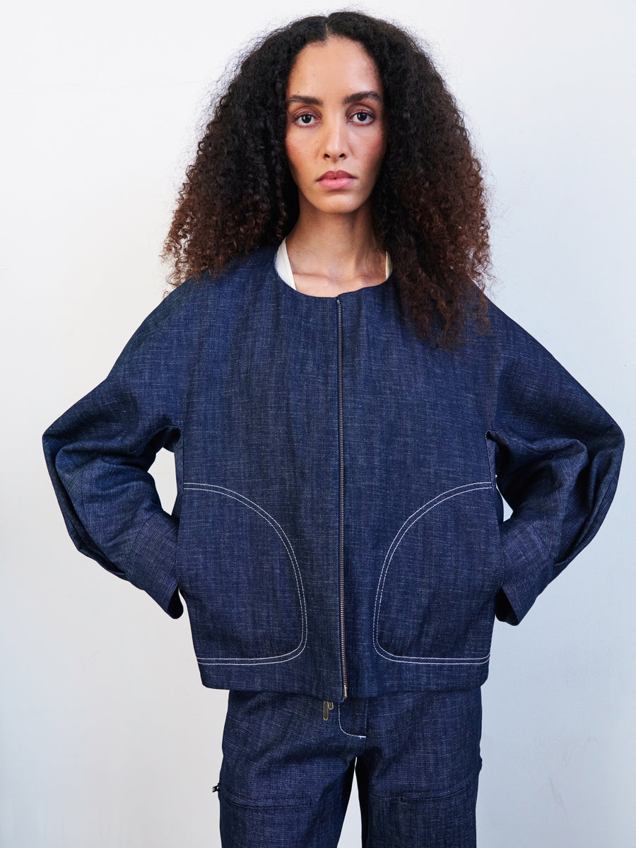 A curly-haired person wears a Zero + Maria Cornejo Akeo Bomber Jacket with matching pants, crafted from dark organic GOTS-certified denim cotton. They pose against a white backdrop, hands on hips, gazing at the camera.