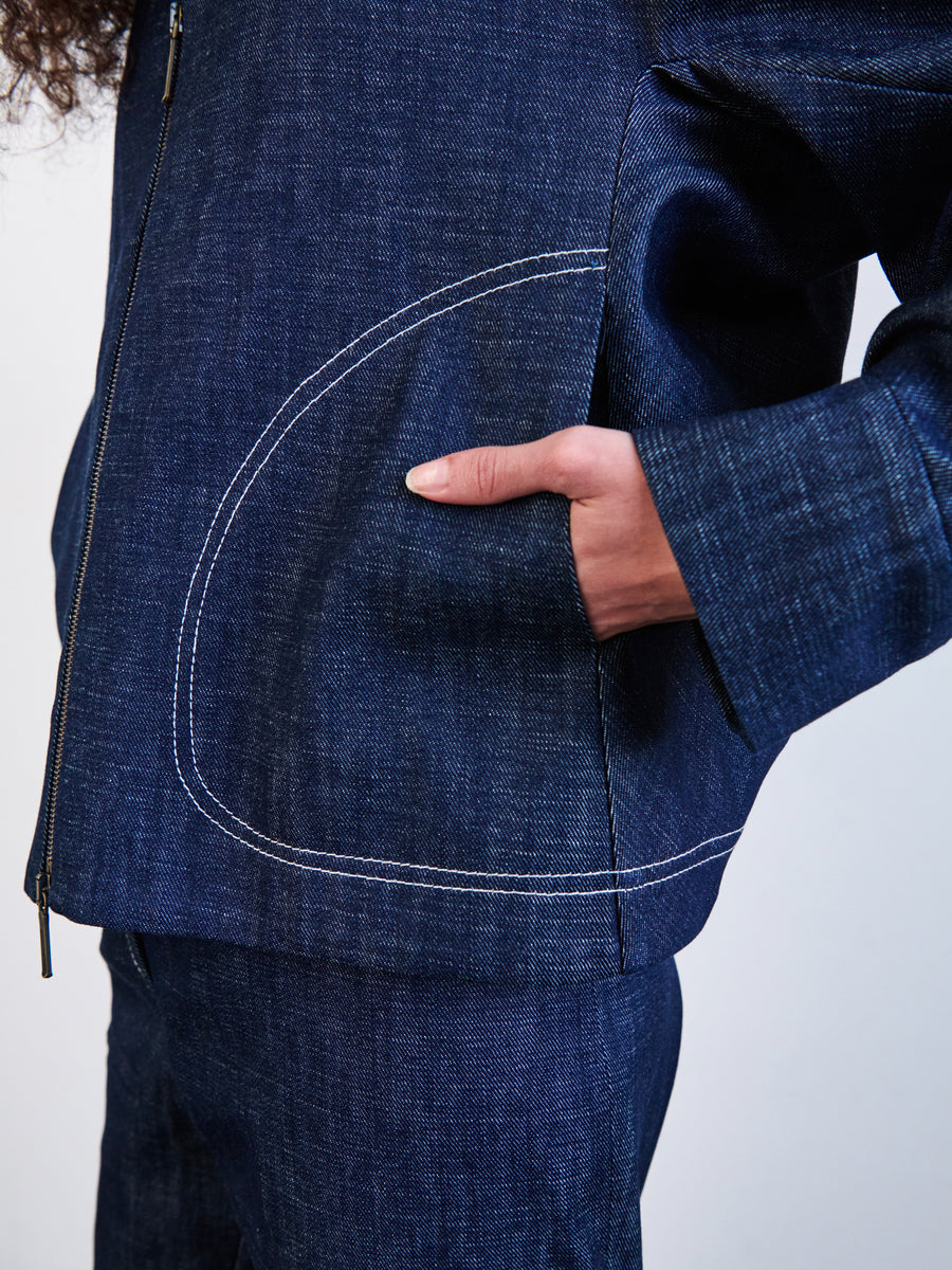 Styled in a New York-made, dark blue Akeo Bomber Jacket by Zero + Maria Cornejo with white stitching and matching pants, a person casually tucks one hand into the large front pocket of the GOTS-certified organic cotton jacket.