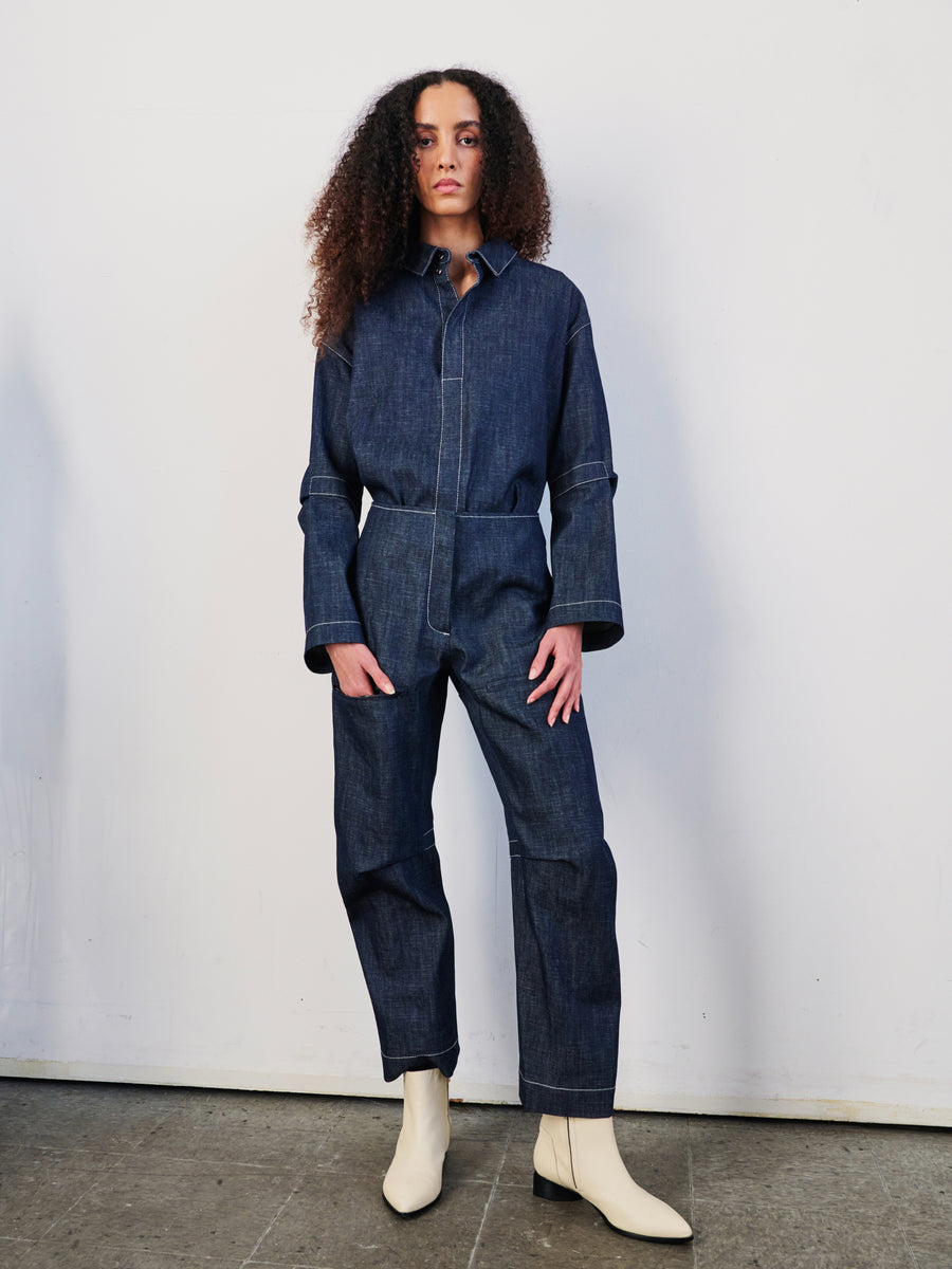 A person with curly hair stands against a plain wall wearing Zero + Maria Cornejo's Denim Biker Pant. The dark denim jumpsuit, crafted from organic denim, features wide cuffs and a relaxed fit. Light-colored boots complete the casual yet stylish look accentuated by the cropped design.