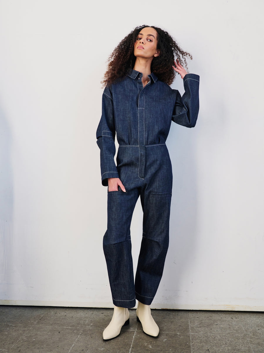 A confident person with curly hair poses against a white wall, wearing Zero + Maria Cornejo's Denim Biker Pant and white ankle boots. The cropped pant accentuates their stance as they rest one hand charmingly on their hair.