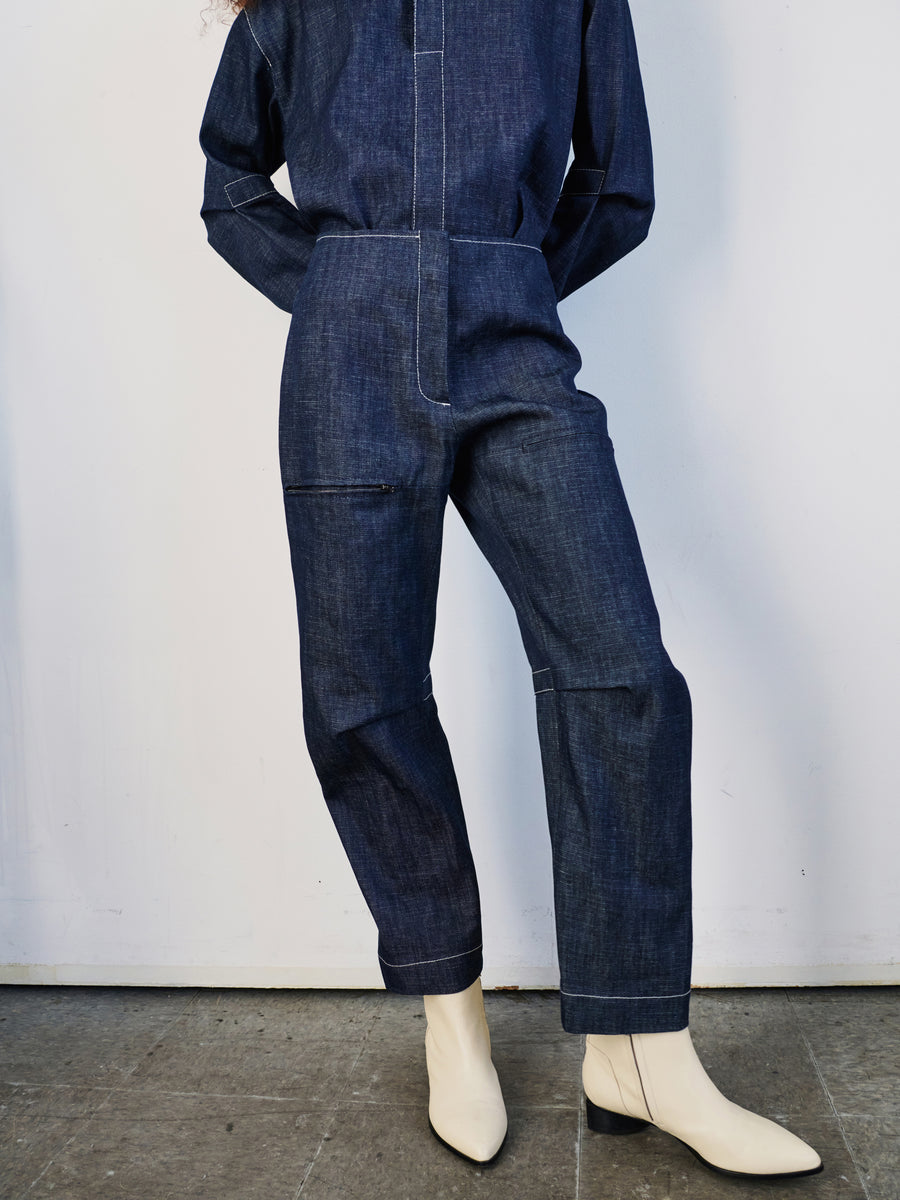 Against a plain white wall, a person poses with hidden hands, wearing the Denim Biker Pant by Zero + Maria Cornejo in dark organic denim and white ankle boots. The pants showcase prominent stitching and a zipper detail on one thigh.