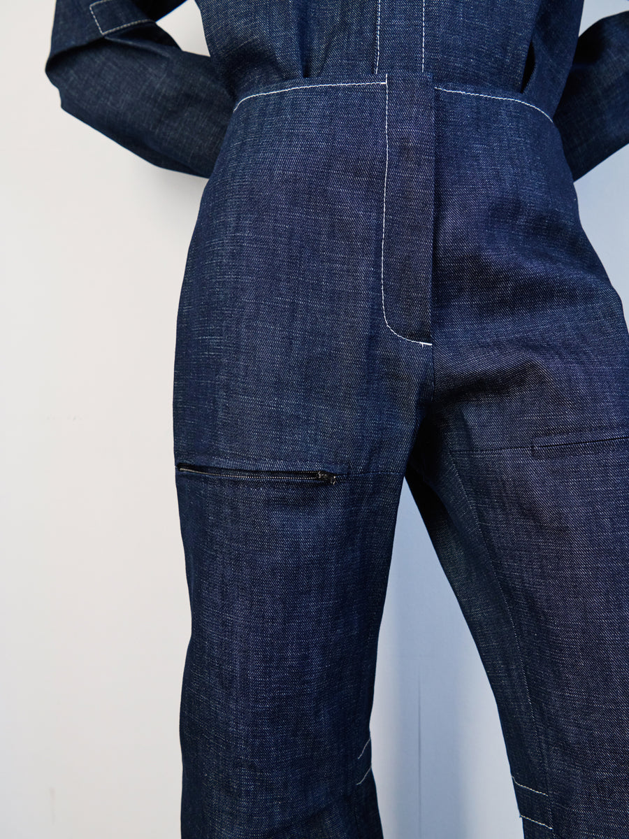 Close-up of someone wearing Zero + Maria Cornejo's Denim Biker Pant in dark blue GOTS-certified organic denim, highlighting visible stitching and a zippered pocket on the pant leg. Hands are behind the back against a plain white background.