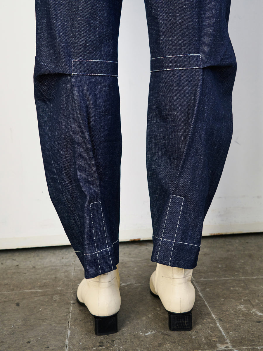 A person stands indoors on a tiled floor, showcasing the rear view of their lower legs in Zero + Maria Cornejo's Denim Biker Pant made from GOTS-certified organic cotton. They pair it with light ankle boots featuring black heels, emphasizing an eco-friendly fashion choice.