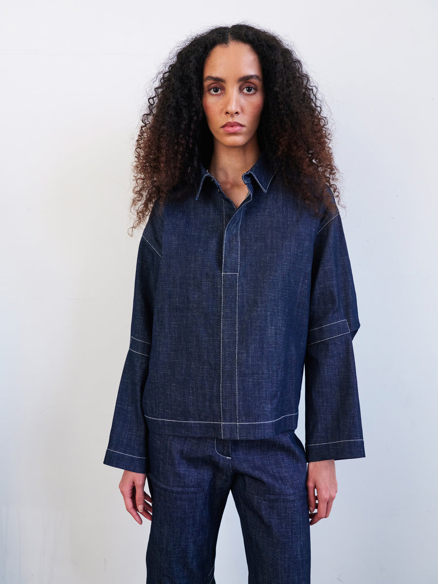 Against a plain white background, a person with long, curly hair wears a dark organic denim outfit by Zero + Maria Cornejo, crafted from GOTS-certified cotton. The Denim Biker Shirt pairs with matching cropped pants featuring visible white stitching.