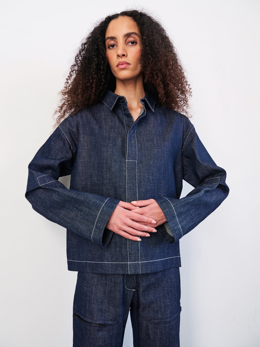 A person with curly hair is wearing a Zero + Maria Cornejo Denim Biker Shirt and GOTS-certified organic denim cropped pants while standing against a plain white background.