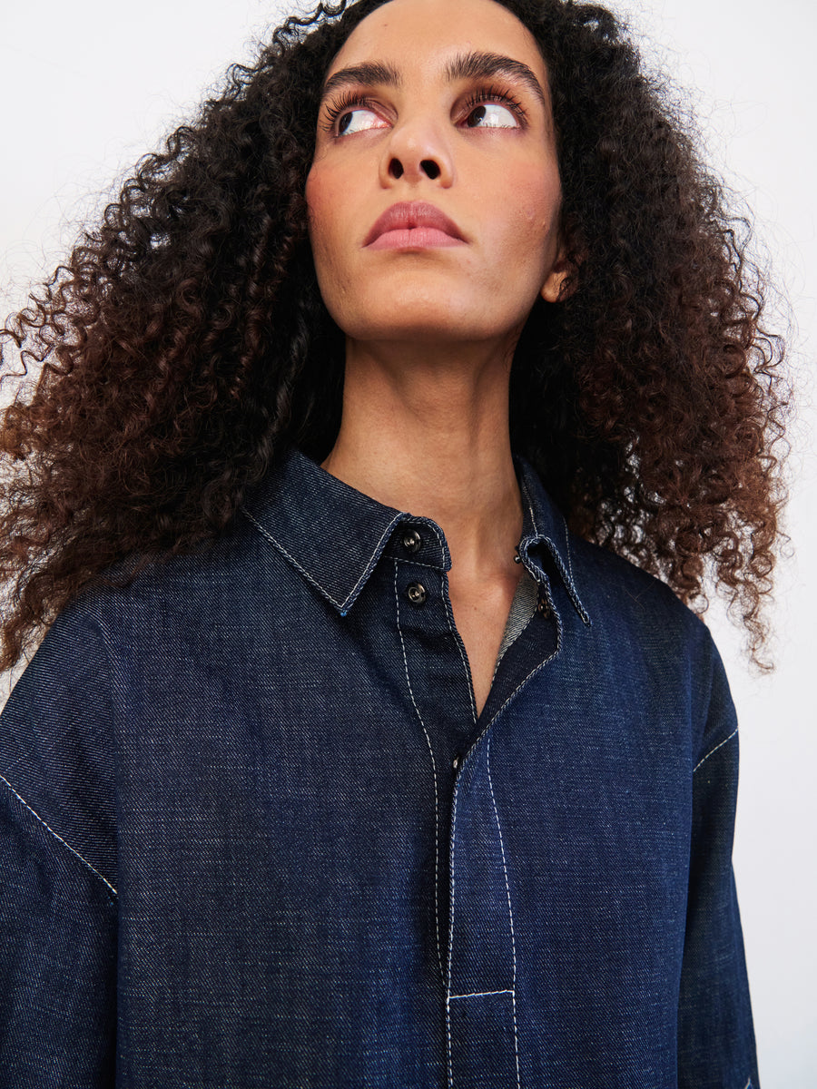 A curly-haired person wears the Denim Biker Shirt by Zero + Maria Cornejo, crafted from GOTS-certified organic denim. The backdrop is a plain, light color, showcasing the shirt's quality as they gaze to the side.