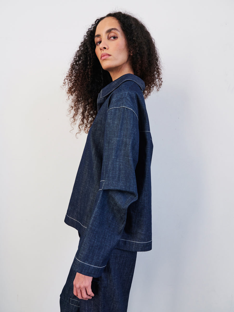 A person with long, curly hair stands sideways in a Zero + Maria Cornejo Denim Biker Shirt and cropped pants against a plain white background, glancing over their shoulder at the camera and exuding effortless style in GOTS-certified cotton attire.