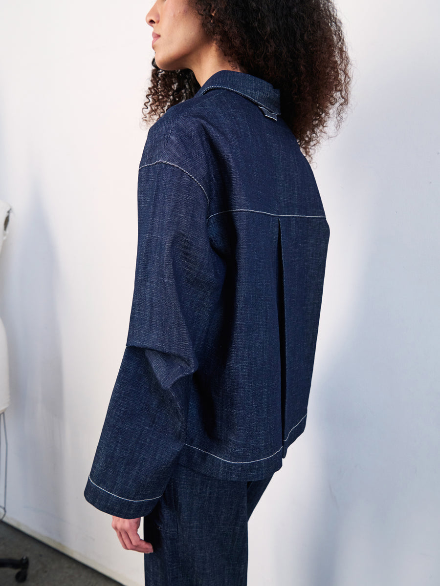 A person with curly hair wears the Denim Biker Shirt by Zero + Maria Cornejo, made from dark organic denim with back pleats and visible seams. They stand sideways against a white wall, completing the look with matching GOTS-certified cotton denim pants.