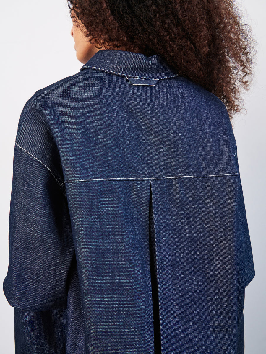 A person with curly hair is shown from the back wearing a Zero + Maria Cornejo Denim Biker Shirt made of GOTS-certified cotton, featuring pleat detailing and visible stitching, paired perfectly with cropped pants. The outfit stands out against a plain white background.