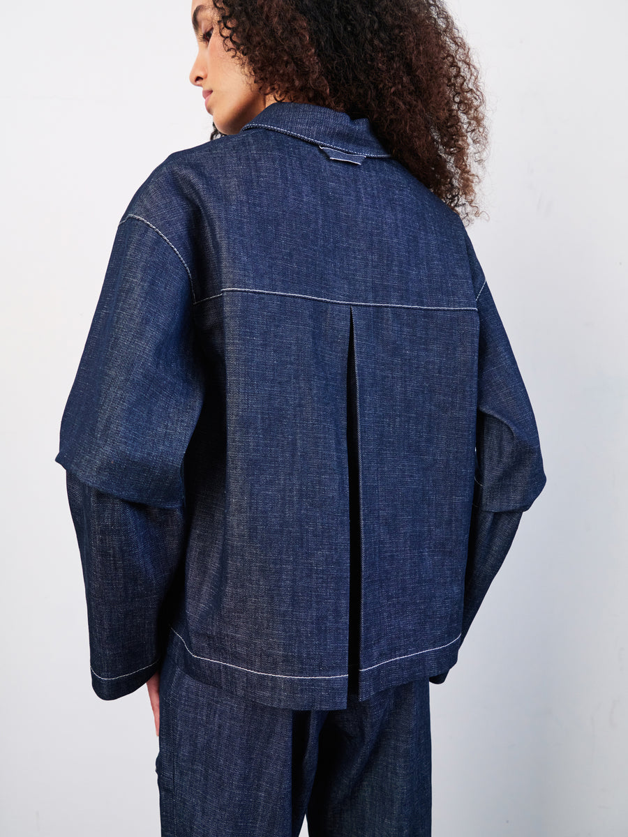 A person with curly hair in a dark denimed Zero + Maria Cornejo Denim Biker Shirt and matching cropped pants is shown from the back. The GOTS-certified cotton outfit boasts white stitching and pleats, set against a plain light backdrop.