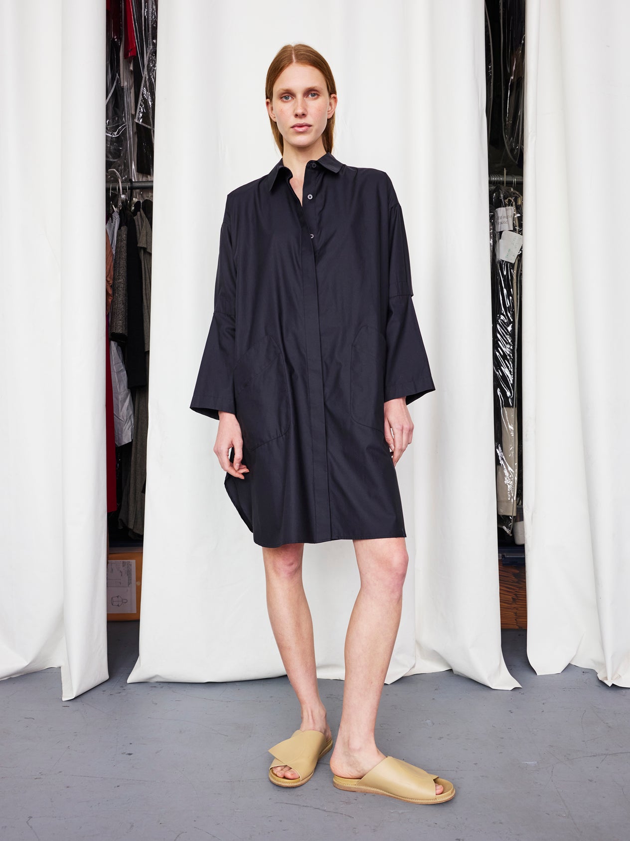 A person stands indoors in the Zero + Maria Cornejo Biker Shirt Dress, a loose-fitting, long-sleeved cotton broadcloth style, paired with beige slide sandals. White curtains and hanging clothes are visible behind them. Machine wash cold for easy care.
