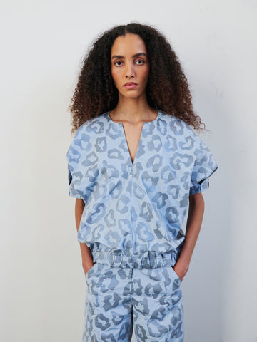 A person with long curly hair wears the Rae Bomber Top, a blue V-neck piece by Zero + Maria Cornejo featuring a metallic tonal animal jacquard pattern and a draped blouson silhouette. They are paired with matching pants made from recycled polyester, standing against a plain white background and facing forward.