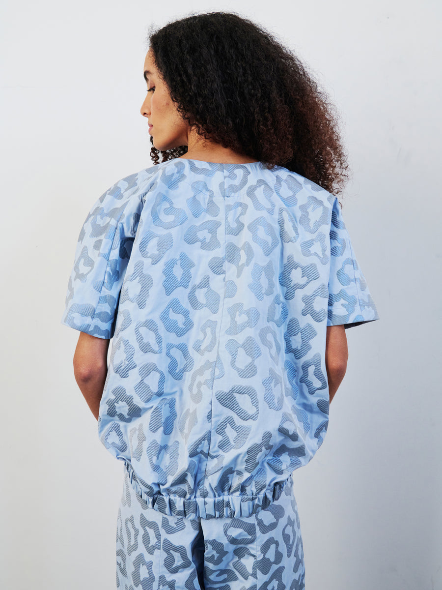 A woman with curly hair is turned to the side, highlighting the Rae Bomber Top by Zero + Maria Cornejo, adorned in large abstract patterns in light blue. The top features a draped blouson silhouette and a cinched waist, crafted from recycled polyester. The image is set against a plain white background.