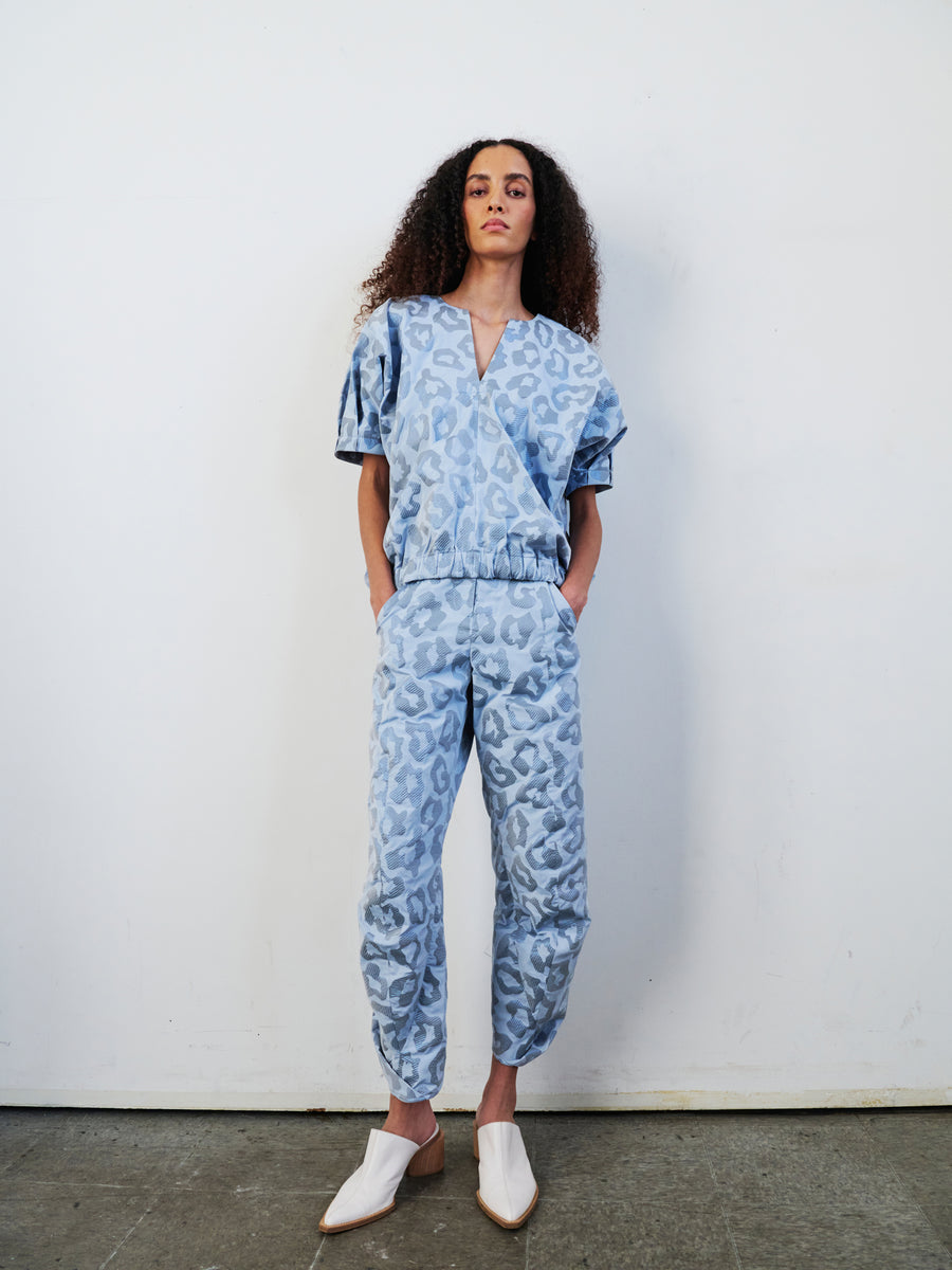 A person with curly hair stands against a white wall dressed in the Takeo Pant by Zero + Maria Cornejo, made from recycled polyester and featuring a blue, leopard-print design. Their white pointed shoes add a sleek touch. Hands in pockets, they maintain a neutral expression, effortlessly blending style and sustainability.