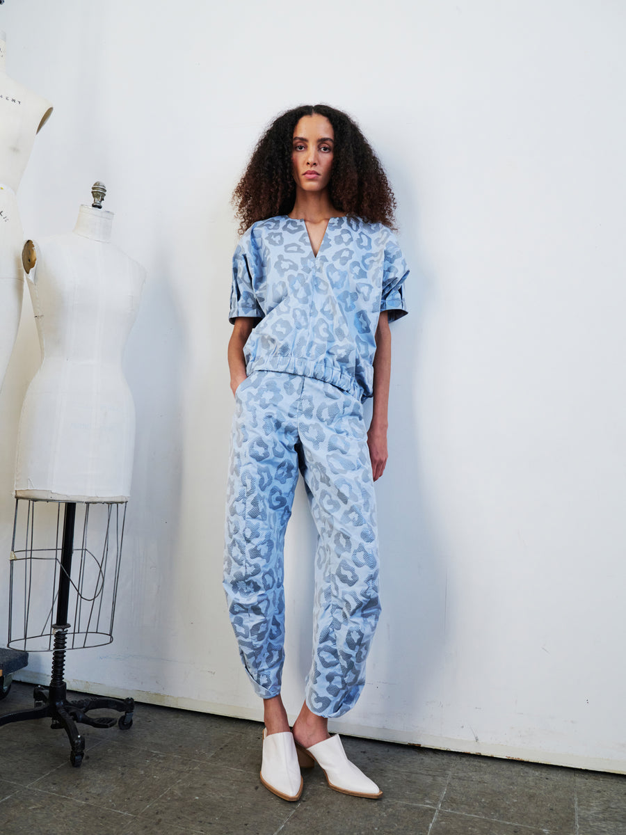 A person with curly hair stands in a room wearing the Takeo Pant by Zero + Maria Cornejo, made from recycled polyester, paired with white shoes. They are next to two dress forms against a plain white wall, looking directly at the camera.