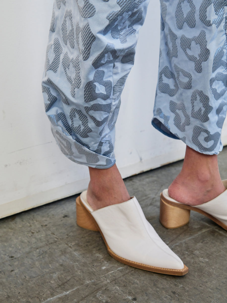 A person wearing white heeled mules is standing on a concrete floor, showcasing the Takeo Pant from Zero + Maria Cornejo. The light blue metallic jacquard pants, featuring a gray abstract pattern, are crafted from recycled polyester and slightly rolled up at the ankles.