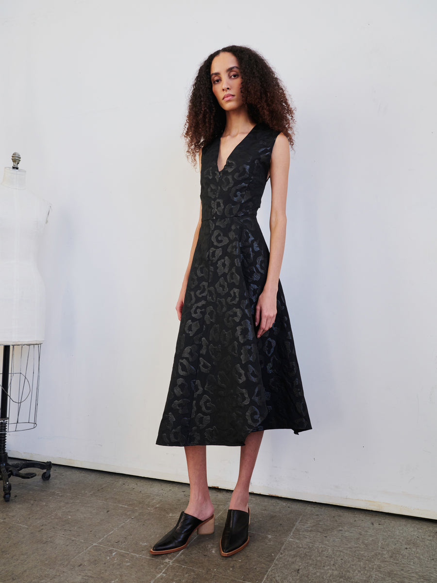 A person with curly hair wears a Sleeveless Wave Dress by Zero + Maria Cornejo and black shoes, standing in a minimalist room with a dress form in the background on the wooden floor.