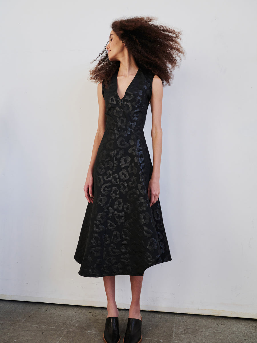 A person with curly hair is wearing a Sleeveless Wave Dress by Zero + Maria Cornejo in black animal jacquard, along with matching shoes. They stand on a concrete floor in front of a plain white wall, looking to their left.