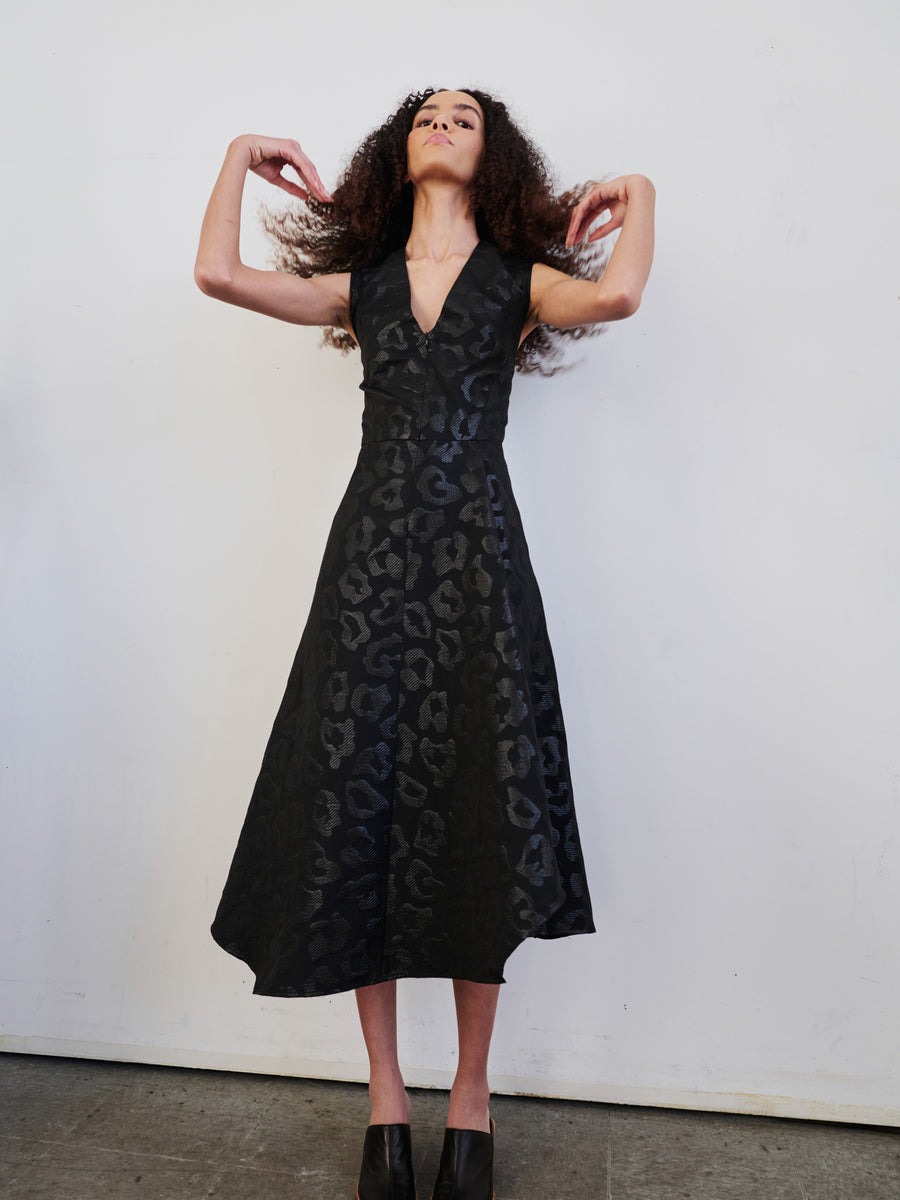 A person stands against a plain wall, wearing a Sleeveless Wave Dress by Zero + Maria Cornejo paired with black shoes. Their curly hair adds flair as they confidently raise their arms, fingers near their hair, creating an expressive and bold pose.