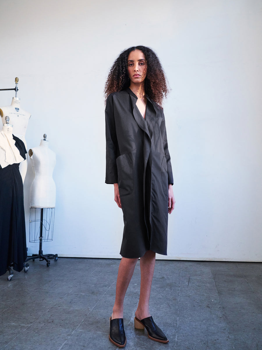 A person with curly hair stands confidently in a minimalist room, wearing the Long-Sleeved Fin Gaban Dress by Zero + Maria Cornejo over slip-on shoes. In the background, tailor mannequins are draped in cotton broadcloth featuring ruffle details.