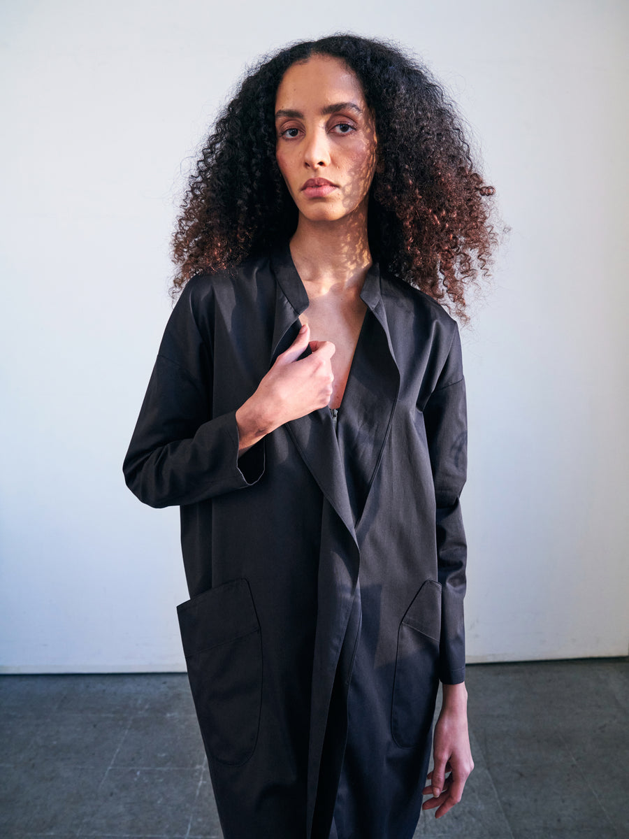 A person with long curly hair wears the fashionable Long-Sleeved Fin Gaban Dress by Zero + Maria Cornejo, featuring ruffle details, as they stand against a plain backdrop. They hold one side of the open dress and gaze directly at the camera in a well-lit room.