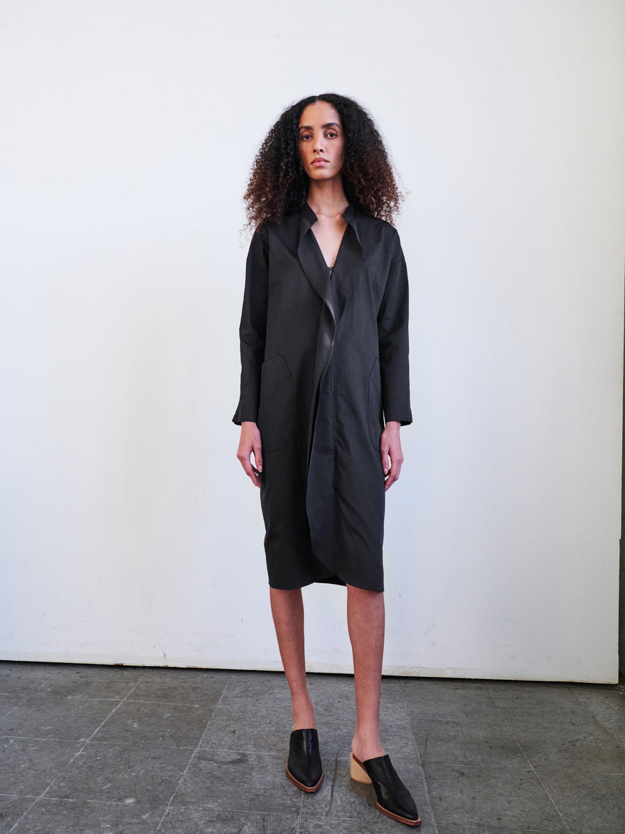 A person with curly hair stands against a plain white background, dressed in the Long-Sleeved Fin Gaban Dress by Zero + Maria Cornejo, which is knee-length and features subtle ruffle detail. They are wearing black slip-on shoes and have a neutral expression while standing on a dark tiled floor.