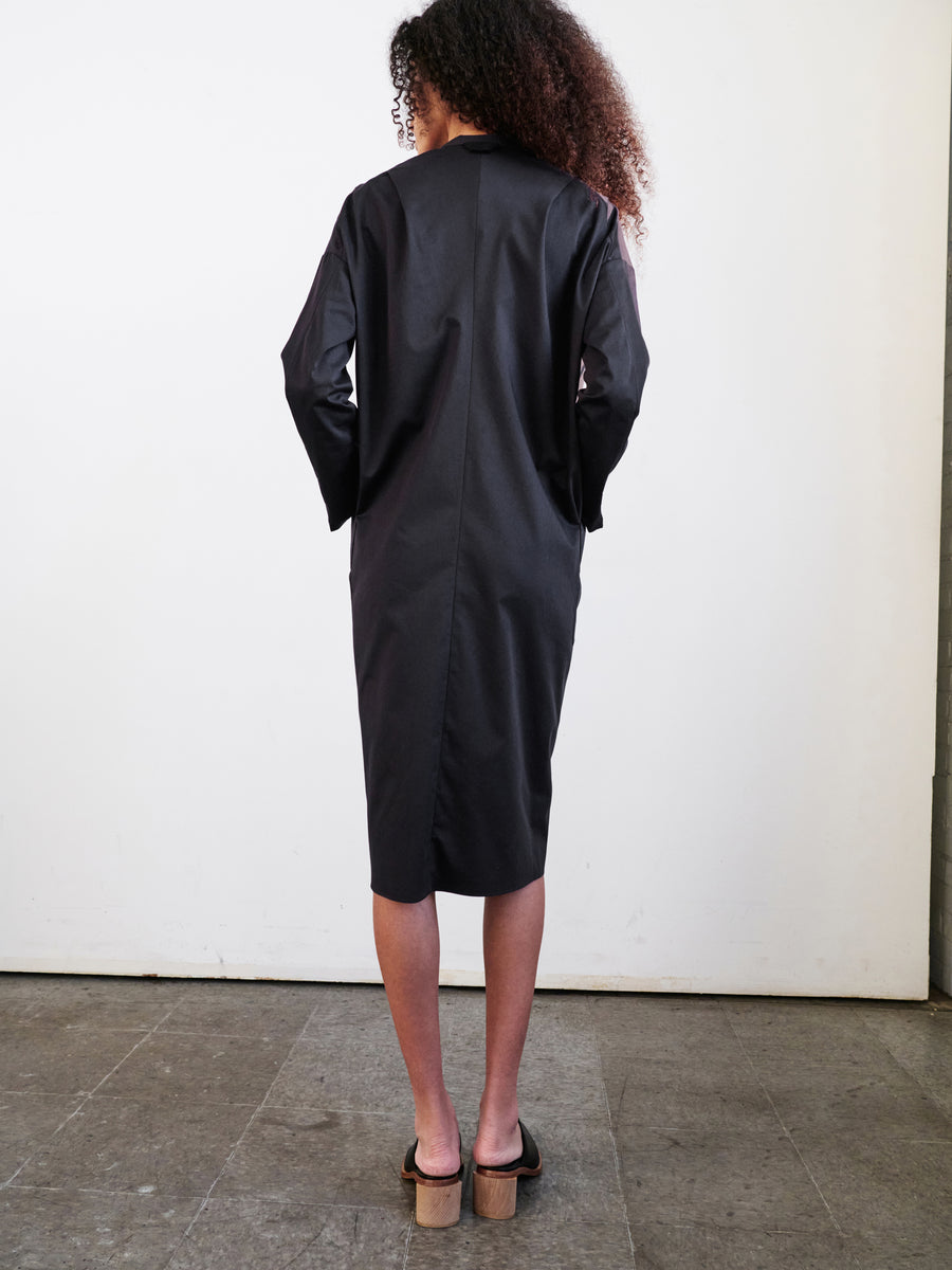 A person with curly hair stands facing a white wall, wearing the knee-length Long-Sleeved Fin Gaban Dress by Zero + Maria Cornejo, paired with sandals. The gray floor contributes to the minimalistic setting.