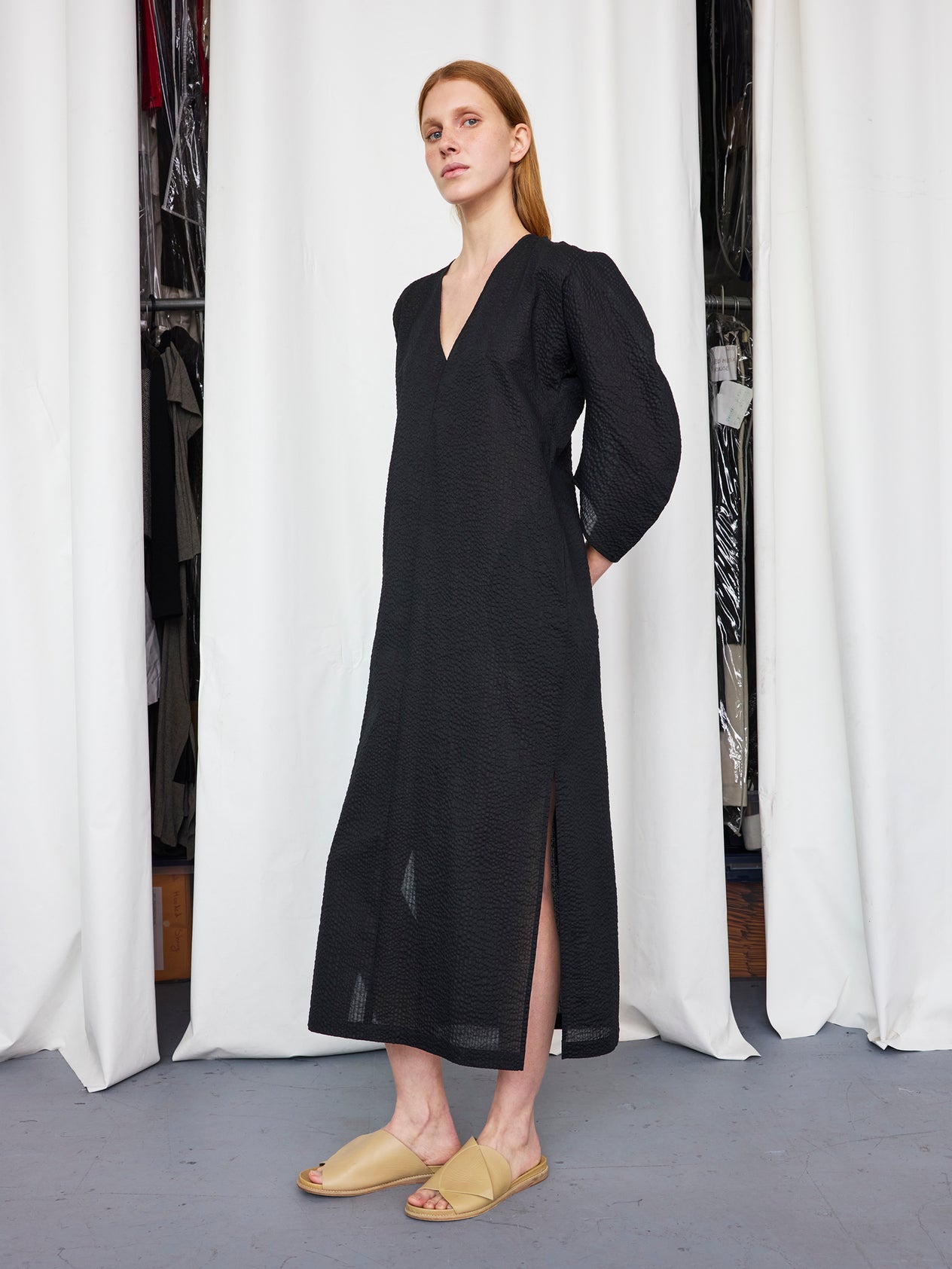 A woman with long, straight hair stands indoors wearing the Zero + Maria Cornejo Long Opi Dress, a loose-fitting black dress with side slits and long sleeves, paired with beige open-toe sandals. White curtains and clothes are in the background.