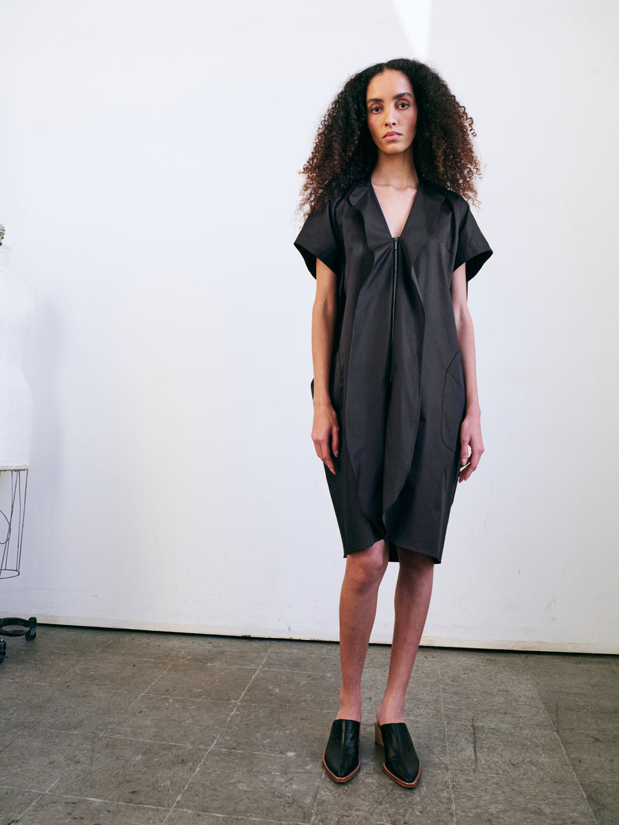 A person with curly hair stands against a plain white wall, wearing the Fin Gaban Dress by Zero + Maria Cornejo, a dark, loose-fitting dress featuring a subtle ruffle detail. They complete the look with black slip-on shoes and are positioned near a dress form on the left.