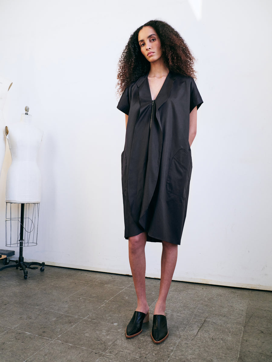 A person with curly hair stands confidently in the Fin Gaban Dress by Zero + Maria Cornejo, a loose, black cotton shirting dress with pockets and a subtly draped collar. They wear black slip-on shoes as a mannequin rests in the background against a plain, light-colored wall.