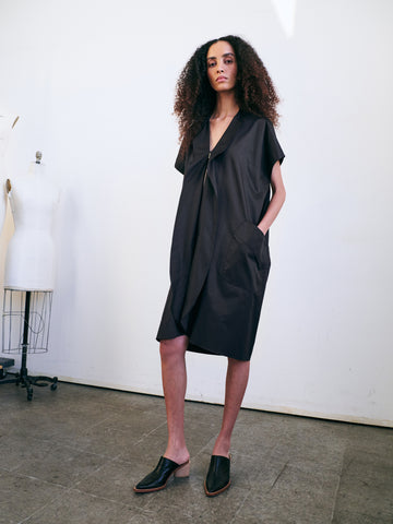 In a minimalist room, a person with curly hair stands confidently wearing the Fin Gaban Dress by Zero + Maria Cornejo, featuring subtle ruffle detail and paired with black slip-on shoes. A dress form is visible in the background.