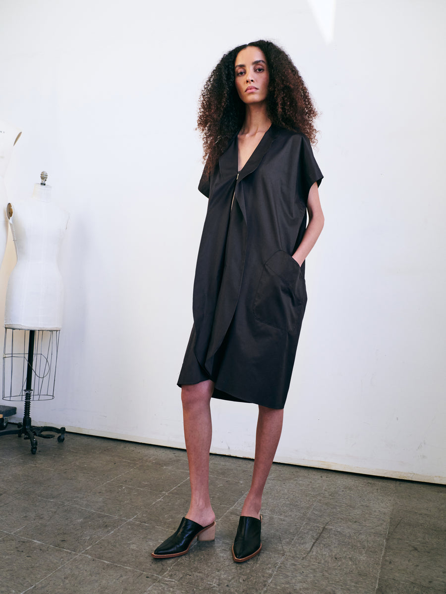 In a minimalist room, a person with curly hair stands confidently wearing the Fin Gaban Dress by Zero + Maria Cornejo, featuring subtle ruffle detail and paired with black slip-on shoes. A dress form is visible in the background.