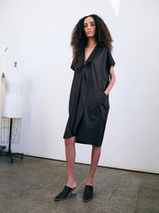 In a minimalist room, a person with curly hair stands confidently wearing the Fin Gaban Dress by Zero + Maria Cornejo, featuring subtle ruffle detail and paired with black slip-on shoes. A dress form is visible in the background.