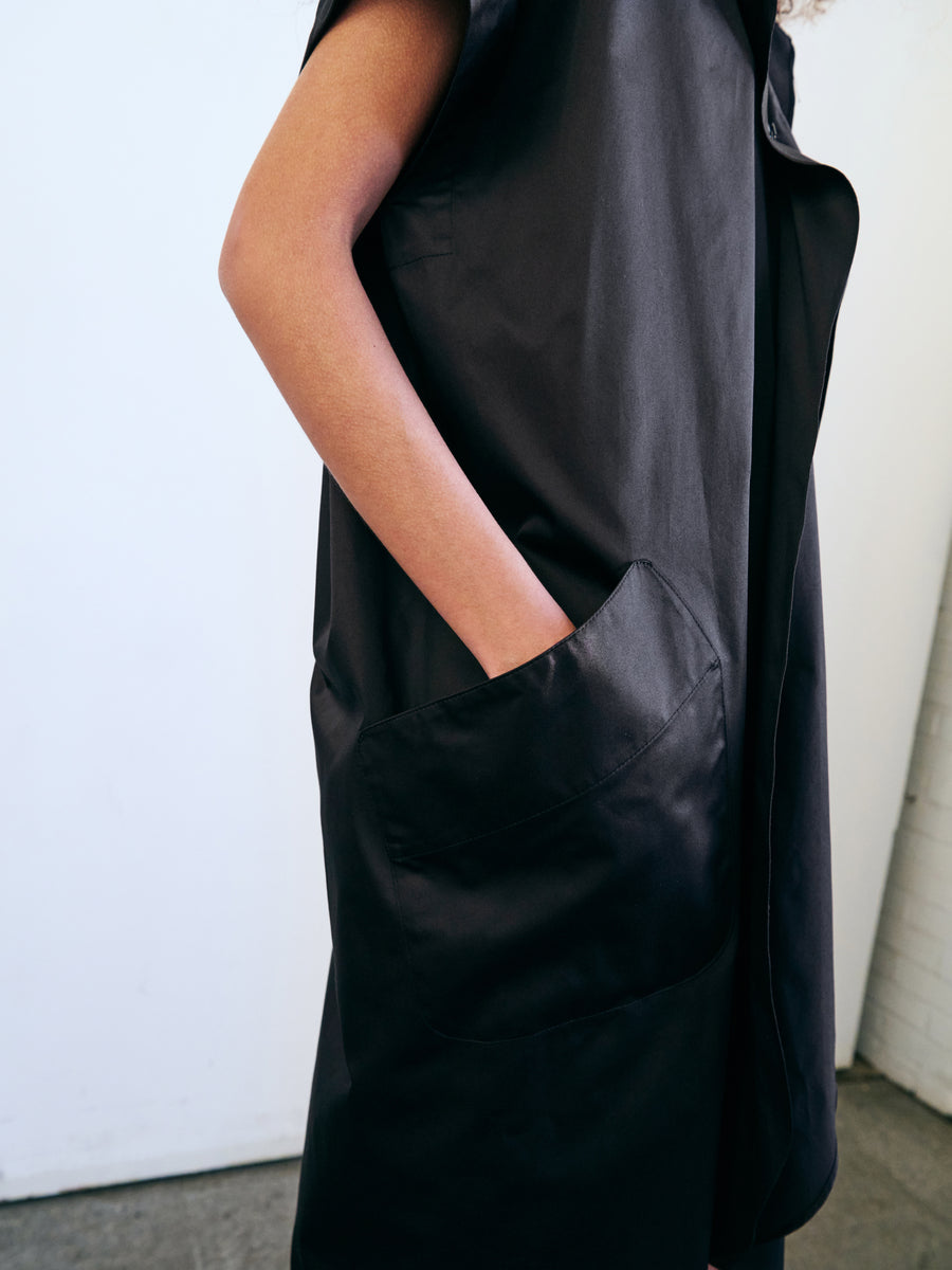 A person is seen wearing the Fin Gaban Dress from Zero + Maria Cornejo, characterized by its sleek black design and sleeveless style with ruffle detail. The dress features a draped collar and a large pocket where the person's hand rests. The smooth texture of the dress is highlighted against a plain white background, and the person's face remains out of view.