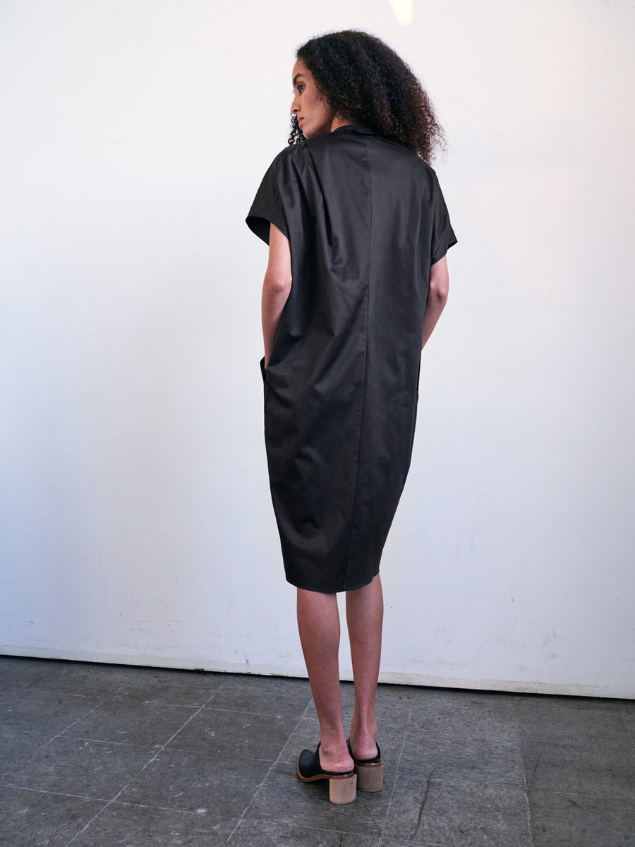 A person with curly hair is standing on a tiled floor, facing a white wall. They are wearing the Fin Gaban Dress – a short-sleeved black cotton shirting dress with subtle ruffle detail by Zero + Maria Cornejo – paired with black slip-on shoes featuring a wooden heel.