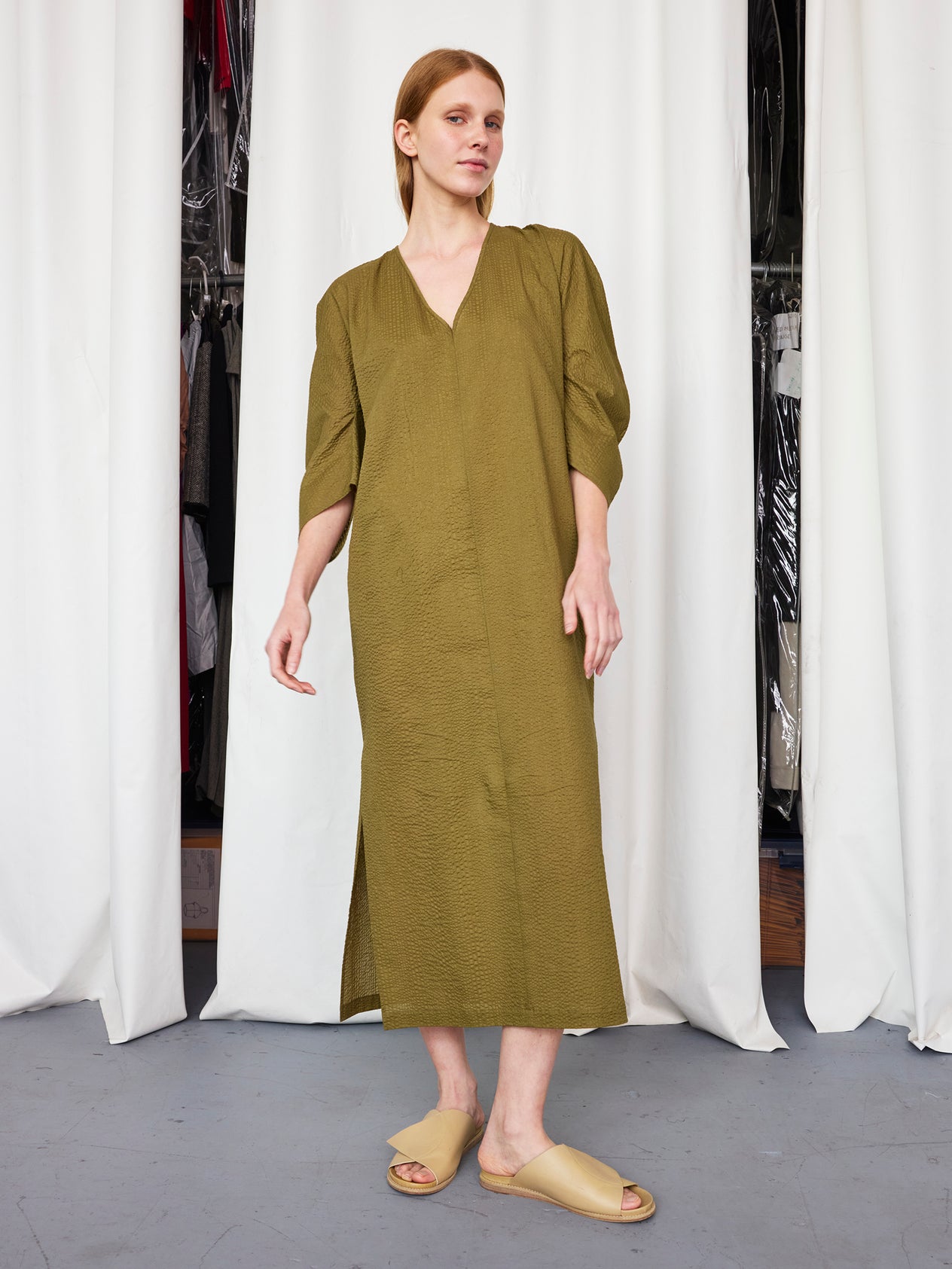 A woman with long, straight hair stands indoors wearing the Zero + Maria Cornejo Long Opi Dress, a loose-fitting black dress with side slits and long sleeves, paired with beige open-toe sandals. White curtains and clothes are in the background.