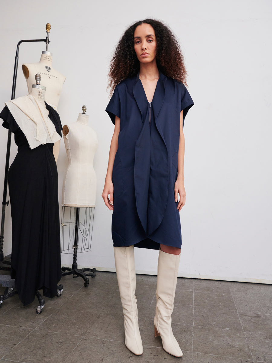 A woman with curly hair stands in a studio wearing the knee-length navy Fin Gaban Dress by Zero + Maria Cornejo, paired with knee-high white boots. Behind her are dress forms, one displaying a black shirting dress and another draped with fabric. The floor is concrete and the walls are white.