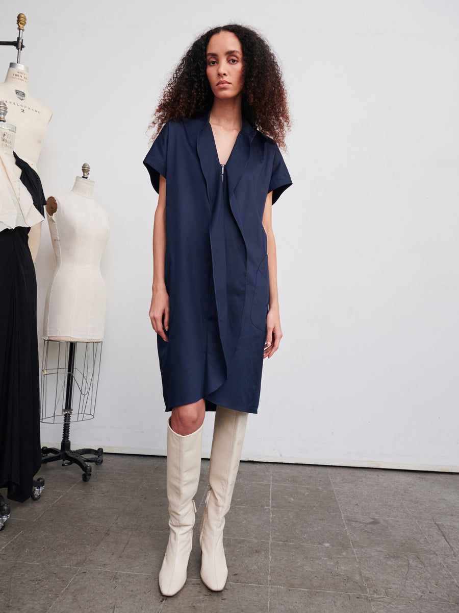 A person with long, curly hair stands in a studio wearing the Fin Gaban Dress by Zero + Maria Cornejo and white knee-high boots. Mannequins with different outfits are in the background against a plain white wall and a gray floor.