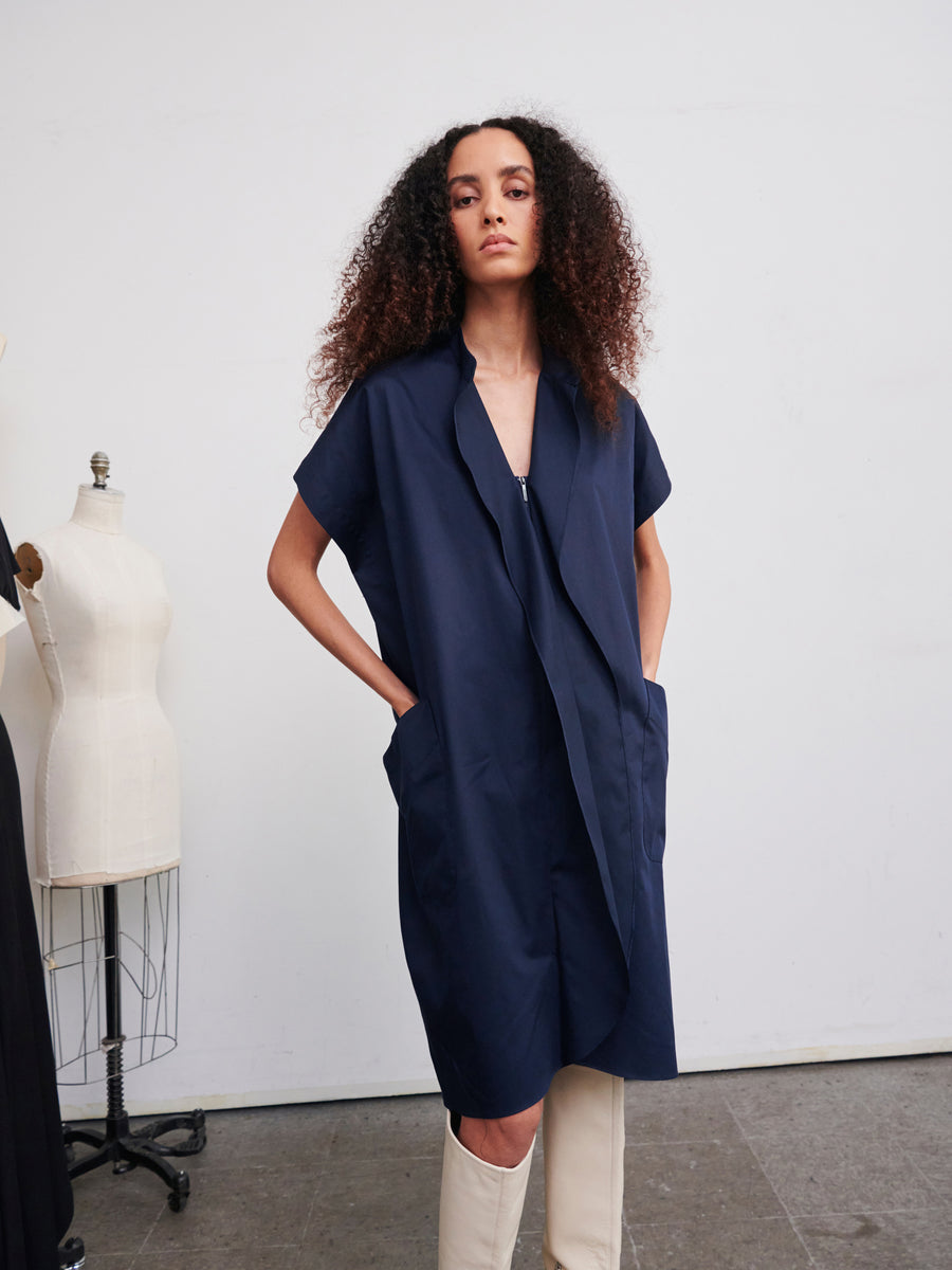 A person with curly hair stands confidently, donning the Fin Gaban Dress by Zero + Maria Cornejo. They are posing in a studio setting, with a dress form visible in the background.