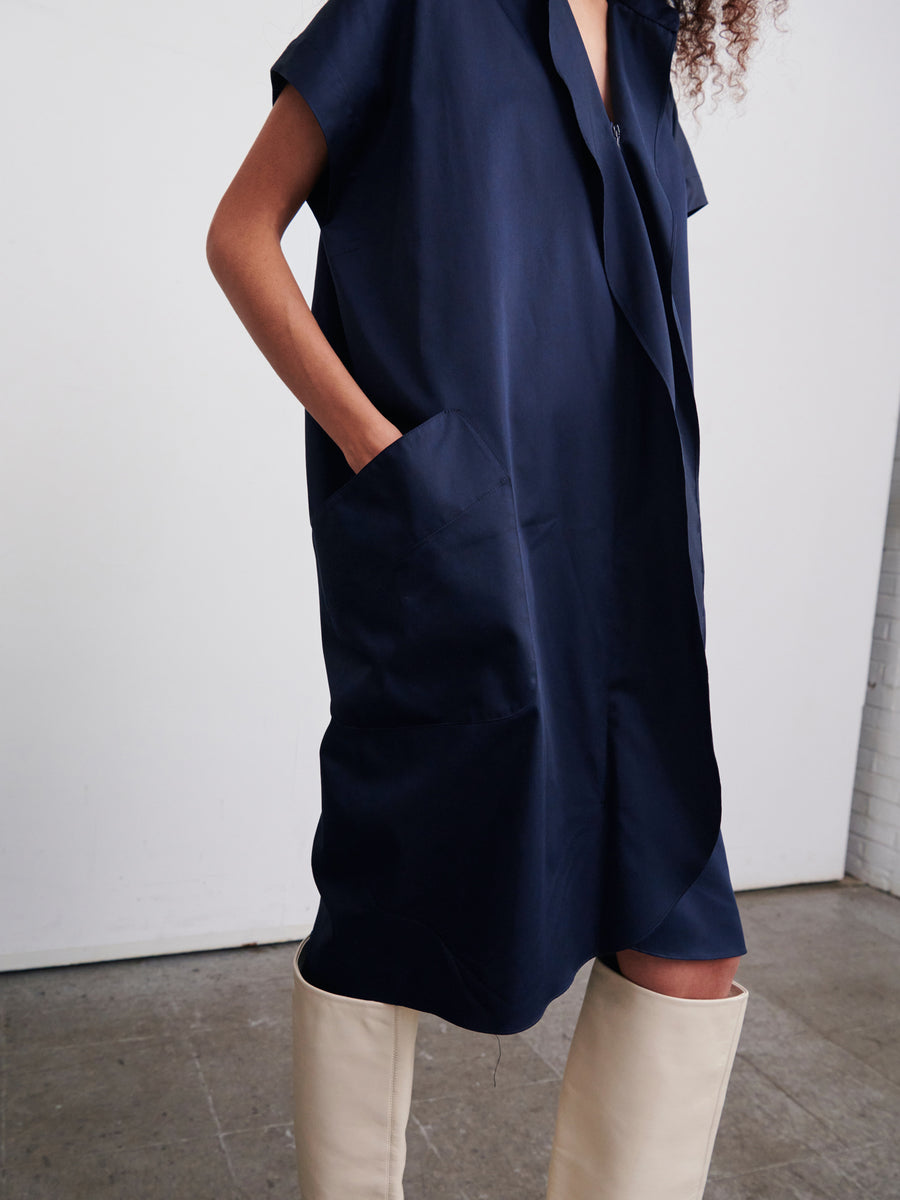 A person wears the Fin Gaban Dress by Zero + Maria Cornejo, a knee-length, loose-fitting dark blue cotton dress featuring short sleeves and convenient pockets. The ensemble is paired with knee-high beige boots set against a plain white wall and gray floor.