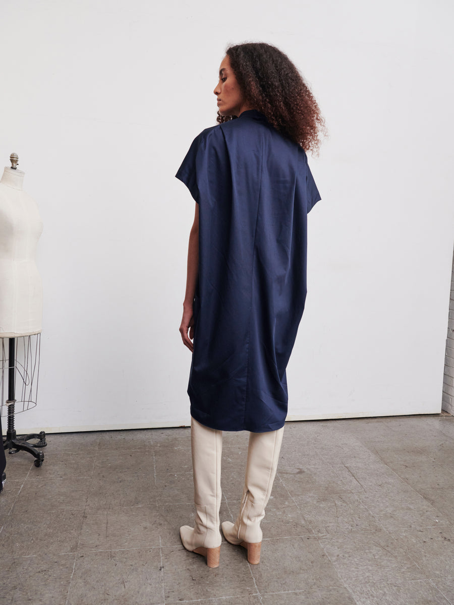 In a minimalist room, a person with curly hair is seen wearing the Fin Gaban Dress by Zero + Maria Cornejo, which is made of navy blue cotton and paired with beige knee-high boots. A dress form can be spotted to the left as the individual faces away, highlighting the back of this knee-length outfit.