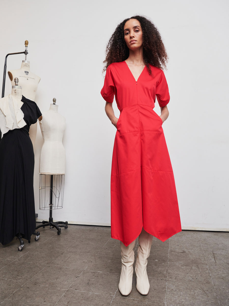 A woman with curly hair is wearing the Aissa Joi Dress in a vibrant red V-neck design by Zero + Maria Cornejo, paired with white boots, as she stands in a fashion studio. She is surrounded by dress forms displaying various garments against a plain, light-colored wall.