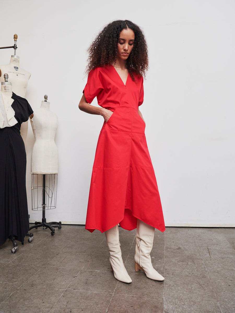 A person with long curly hair is wearing the vibrant red Aissa Joi Dress from Zero + Maria Cornejo and white knee-high boots, posing in a fashion studio with mannequins in the background.