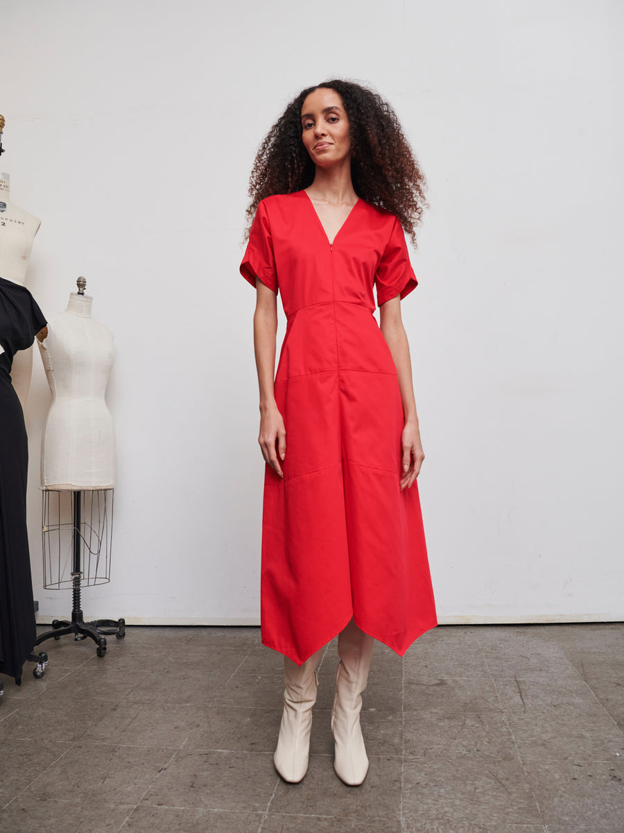 A woman with curly hair wears the vibrant red Aissa Joi Dress by Zero + Maria Cornejo and pairs it with white boots, standing confidently in a minimalist studio. The A-line skirt flows elegantly as two dress forms linger subtly in the background.