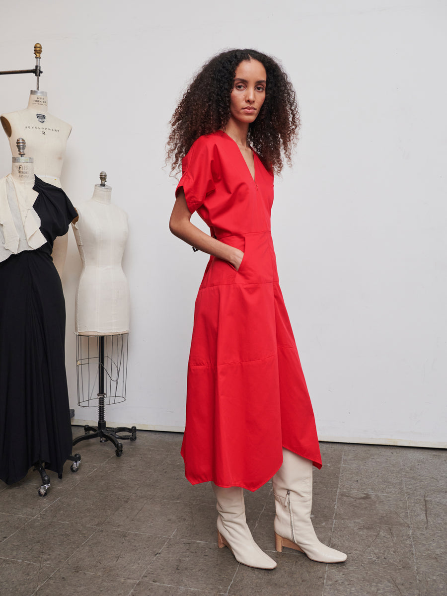 A person with long curly hair is wearing the striking bright red Aissa Joi Dress by Zero + Maria Cornejo, paired with white boots, standing in a studio with mannequins in the background. The setting suggests a vibrant fashion design environment.