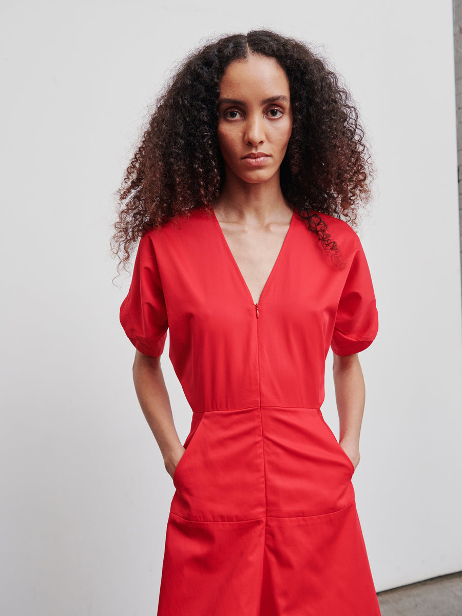 A person with long, curly hair wears the Aissa Joi Dress by Zero + Maria Cornejo, featuring a bright red cotton broadcloth design with an A-line skirt, V-neck, and short sleeves. They stand against a plain white background, looking directly at the camera with their hands in the dress pockets.