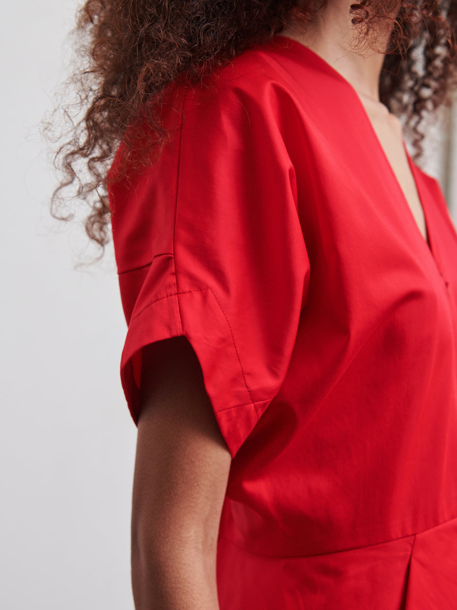 A person with curly hair is wearing the Aissa Joi Dress by Zero + Maria Cornejo, a vibrant red cotton broadcloth dress. The focus is on the upper half, highlighting its smooth fabric and V-neck design, while the person's face remains out of view.