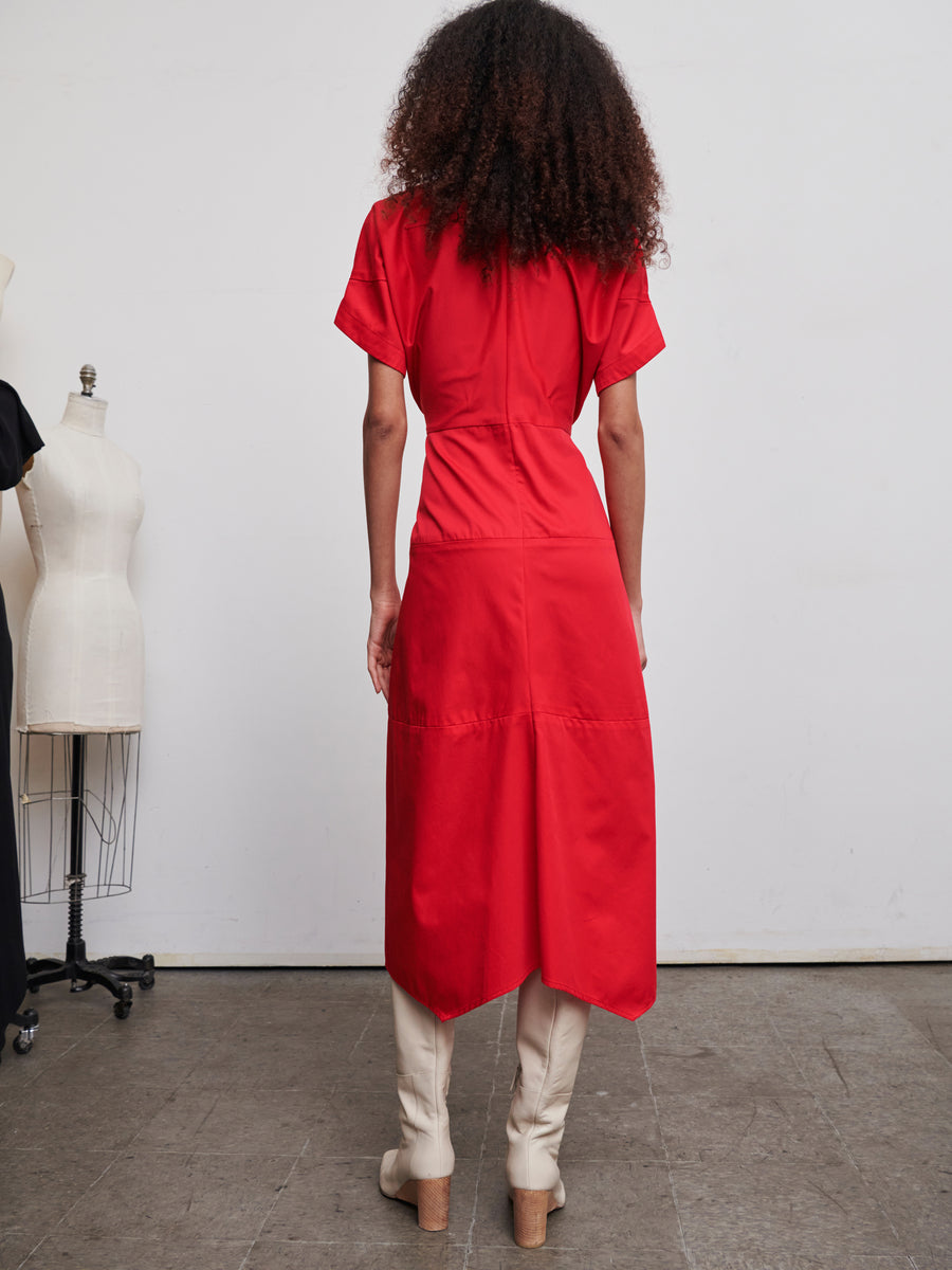 A woman with curly hair stands with her back to the camera, wearing the vibrant red Aissa Joi Dress from Zero + Maria Cornejo and cream-colored boots. She is in a studio setting adorned with dress mannequins in the background.